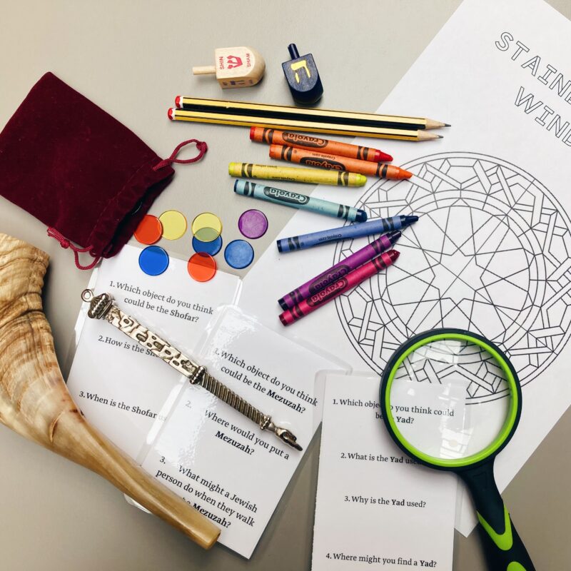 The image shows a variety of educational and cultural items laid out on a table. There are two dreidels, a Shofar (ram's horn), a Mezuzah, a magnifying glass, crayons, colored pencils, circular colored chips, and a red velvet drawstring bag. Additionally, there are worksheets with questions related to Jewish objects and practices, including a coloring page with a stained glass window design.