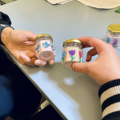 Two hands holding small candle jars with coluorful designs, made during an arts activity at the museum.