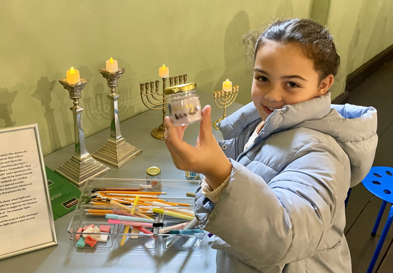 A young girl in a grey jacket holding a small candle jar which she decorated during an arts activity at the museum. 