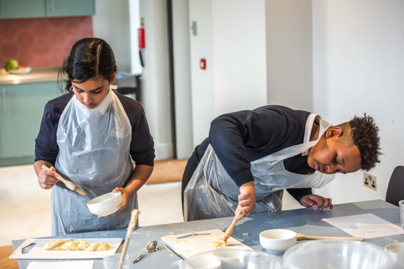 Two young students are engaged in a baking activity in a modern kitchen setting. Both are wearing dark clothing covered by clear plastic aprons. The student on the left is brushing a baking tray of dough with an egg wash from a small bowl, while the student on the right is leaning over the table, focusing intently on their task with a wooden brush. Various baking utensils and ingredients are scattered on the table. The kitchen background includes green cabinets and a red wall, creating a vibrant environment.