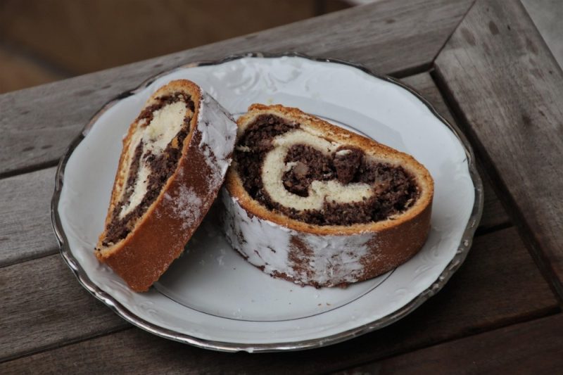 Two slices of a swirled poppy seed roll cake on a decorative white plate, dusted with powdered sugar, sitting on a weathered wooden table.