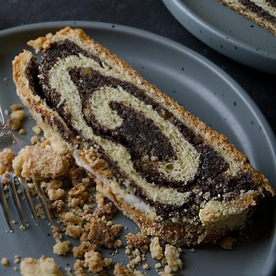 Two gray plates with slices of poppy seed roll, showing the swirled poppy seed filling and crumb topping. A tin of chocolate-dipped cookies is visible in the upper left corner.